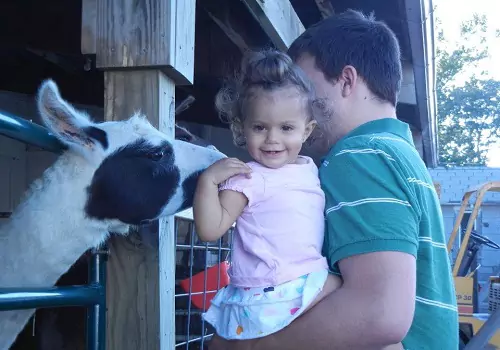 A father holds his daughter while a goat nuzzles up against her at Tanners Orchard, which offers one of the Best Ways to Celebrate Your Child's Birthday.