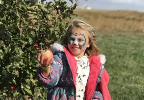A girl with a painted face smiles and shoves an apple toward the camera. There are many Health Benefits of Apples.
